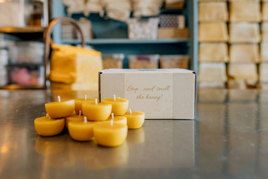 Pure beeswax tea light candles stacked on workshop table with "Stop and smell the honey" gift box and shelves of handcrafted candles in background